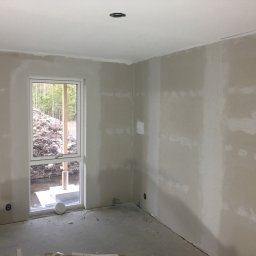 Dawid Fornal - Wnętrze w trakcie prac wykończeniowych: ściany po szpachlowaniu, widoczne nierówności i ślady po łatach, surowy beton na podłodze, okno z widokiem na ziemię i drzewa.