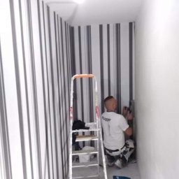 Dawid Fornal - Pracownik w koszulce firmy Flugger szpachluje ścianę w wąskim pomieszczeniu z tapetą w czarno-białe pionowe pasy, obok stoi drabina.
