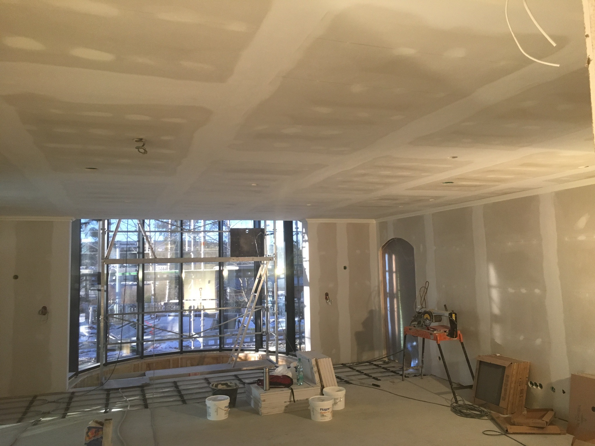 Wnętrze w trakcie prac wykończeniowych: surowy sufit z widocznymi łączeniami płyt gipsowo-kartonowych, ściany przygotowane do dalszych prac, duże okno z widokiem na zewnątrz, narzędzia i materiały...