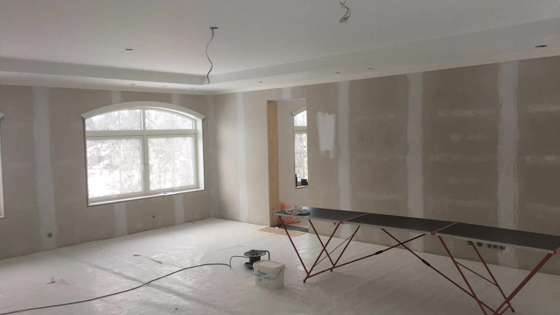 Wnętrze w trakcie szpachlowania: ściany z widocznymi łączeniami płyt gipsowo-kartonowych, zabezpieczona podłoga, rozłożony stół roboczy, wiadro z masą szpachlową Flugger.