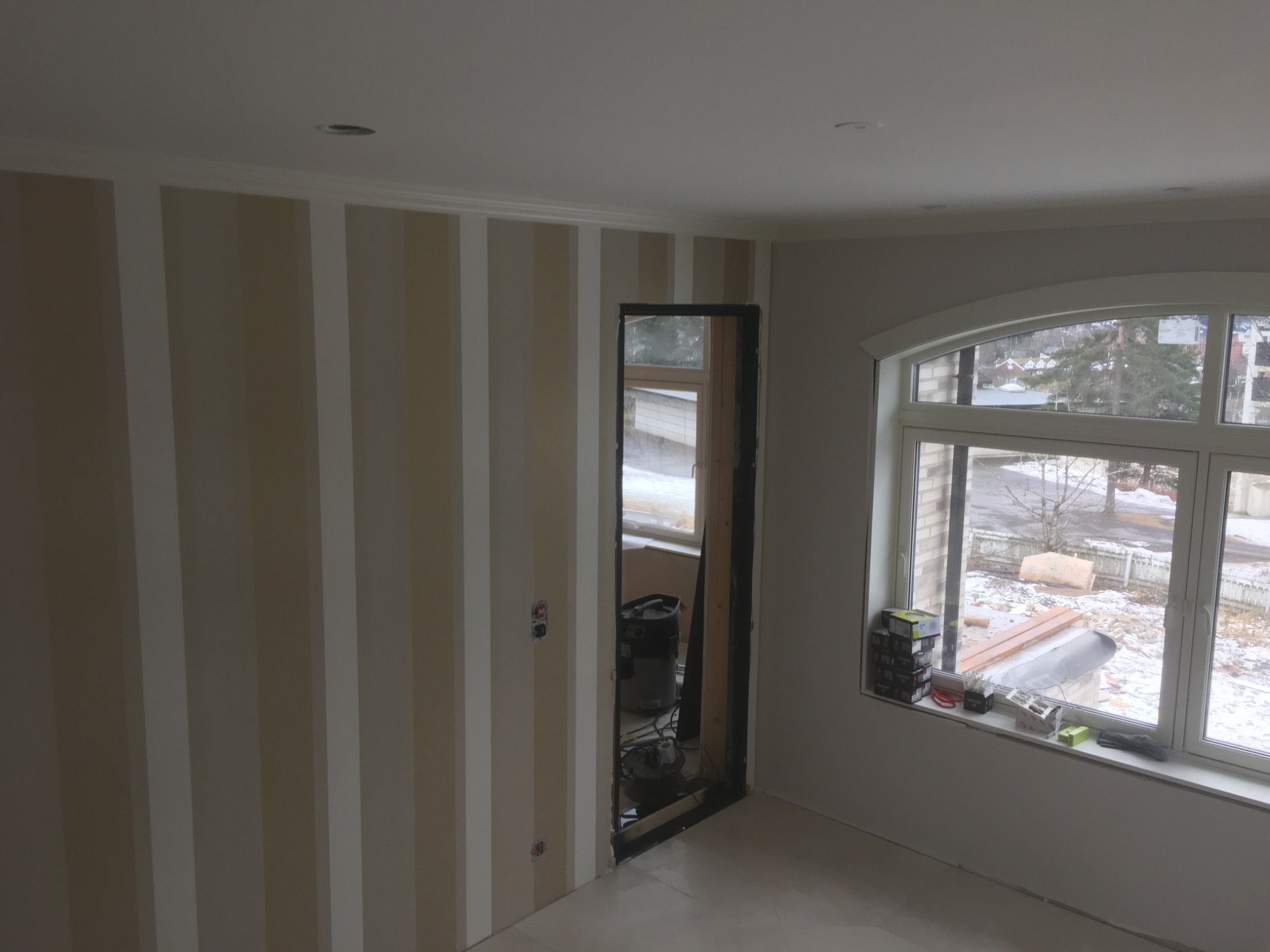 Pokój w trakcie prac wykończeniowych: ściana w pionowe pasy, otwór drzwiowy bez drzwi i okno z widokiem na zimowy krajobraz z narzędziami na parapecie.