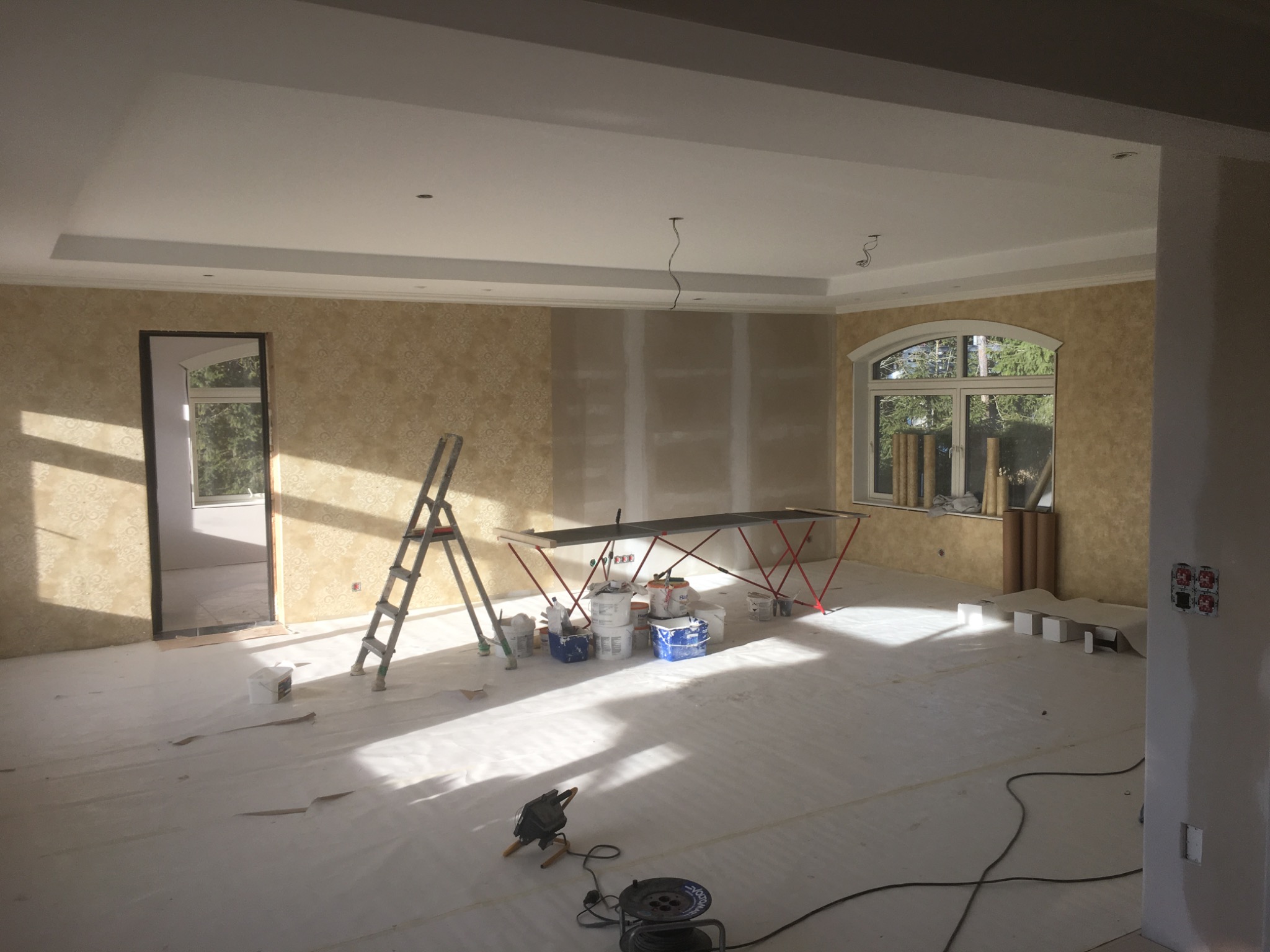 Pokój w trakcie remontu: ściany częściowo z tapetą w kwiaty, częściowo przygotowane do szpachlowania, podłoga zabezpieczona folią, widoczne drabina, farby, narzędzia malarskie i oświetlenie...