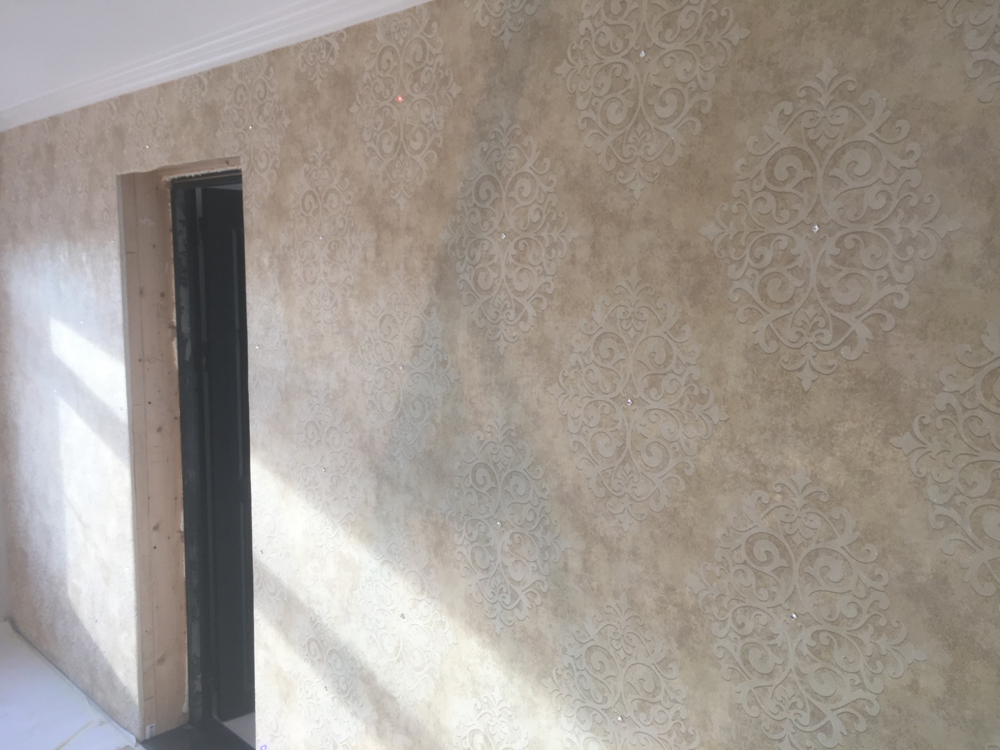Ściana z dekoracyjną, beżową tapetą w ornamenty, częściowo oświetlona przez promienie słoneczne wpadające przez okno. Widoczny fragment ościeżnicy drzwiowej w trakcie montażu.