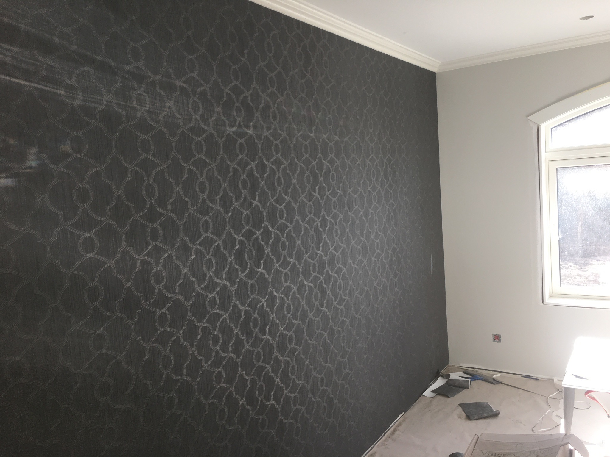 Ściana pokryta ciemnoszarą tapetą z delikatnym, geometrycznym wzorem, widoczna listwa przypodłogowa i fragment okna, przygotowania do dalszych prac wykończeniowych.