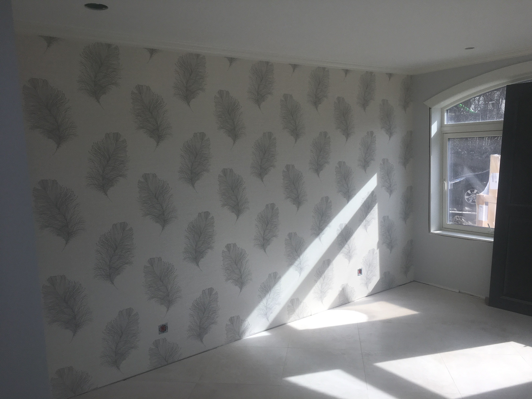 Jasne pomieszczenie po malowaniu ścian, pokryte tapetą w szare piórka, z widocznym oknem i padającym światłem słonecznym.