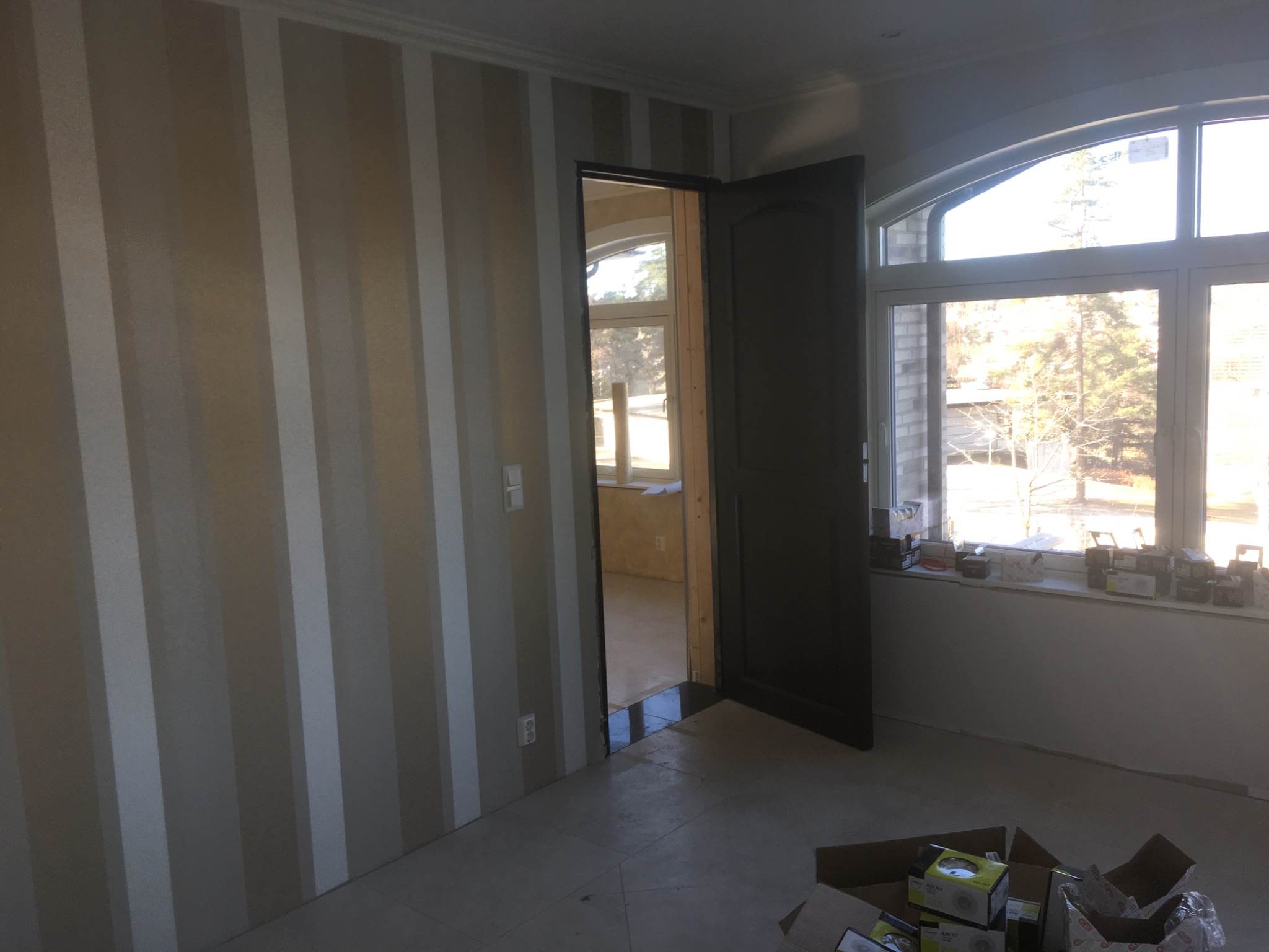 Pokój po remoncie: ściana w pionowe pasy beżu i bieli, otwarte ciemne drzwi, okno z widokiem na zewnątrz, kartony z akcesoriami na podłodze.