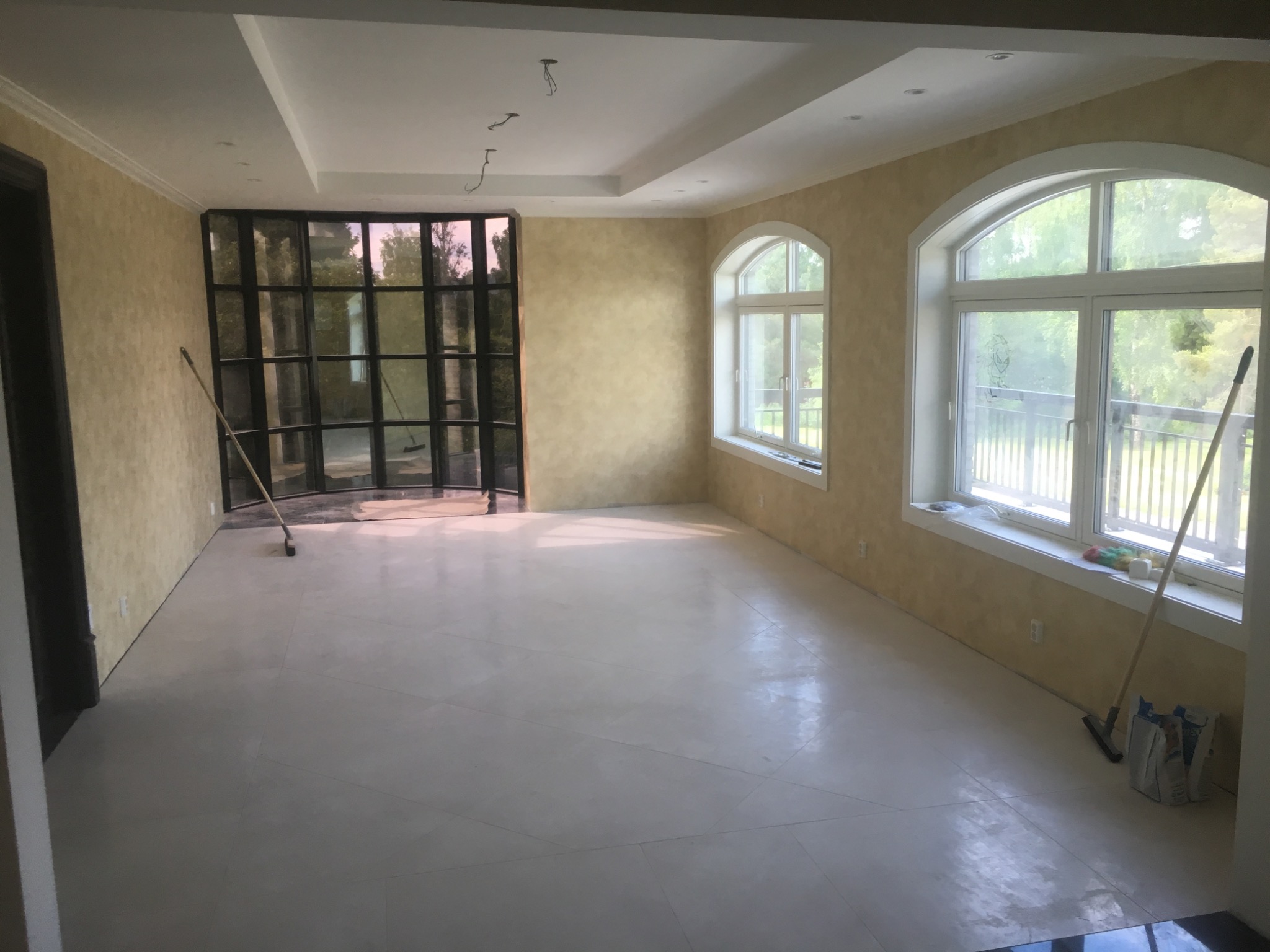Przestronne wnętrze z dużymi oknami, świeżo wykończonymi ścianami w odcieniach beżu i jasną podłogą z płytek; widoczne ślady po remoncie, takie jak szczotki i worki przy oknie.