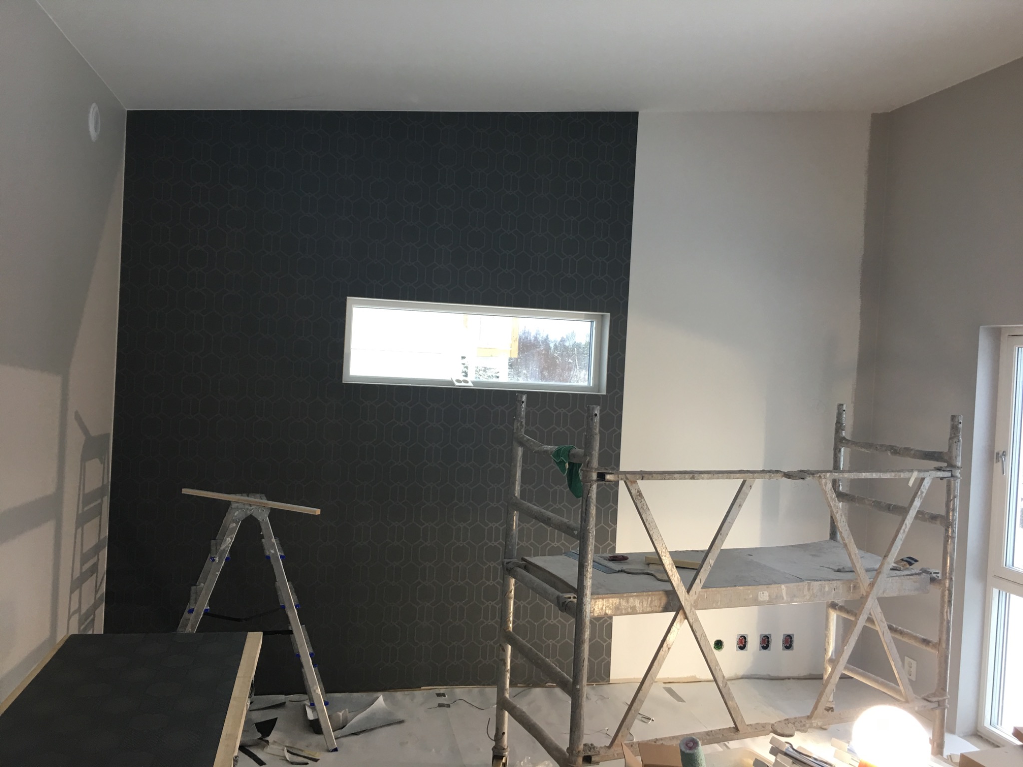 Pokój w trakcie remontu: ściana pokryta ciemną tapetą z geometrycznym wzorem, obok ściana pomalowana na biało, widoczne rusztowanie i drabina malarska, podłoga zabezpieczona folią malarską.