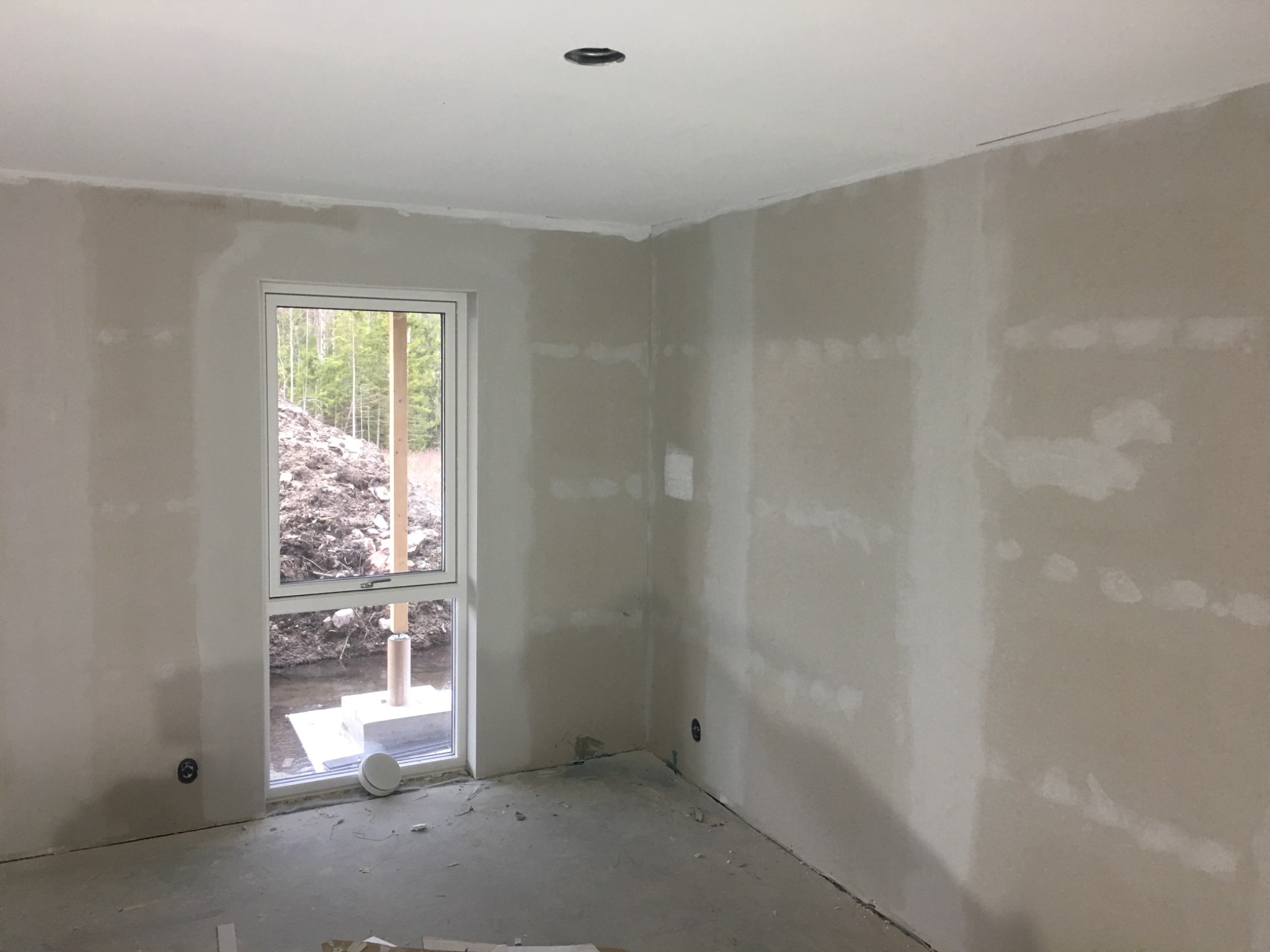 Wnętrze w trakcie prac wykończeniowych: ściany po szpachlowaniu, widoczne nierówności i ślady po łatach, surowy beton na podłodze, okno z widokiem na ziemię i drzewa.