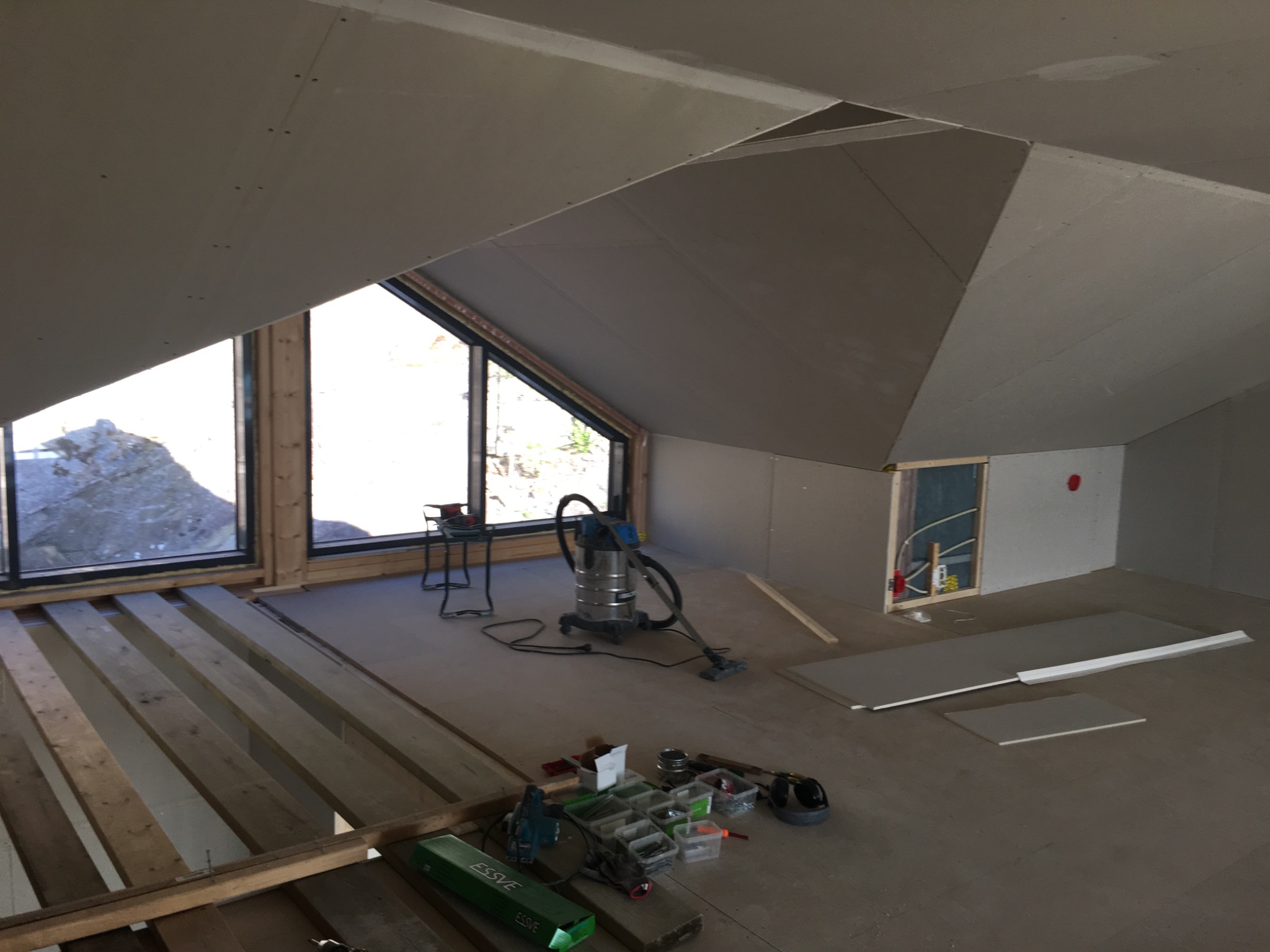 Poddasze w trakcie prac wykończeniowych we Wrocławiu: widoczne płyty gipsowo-kartonowe na ścianach i skośnym suficie, odsłonięta konstrukcja podłogi, okna dachowe oraz narzędzia i odkurzacz budowlany.