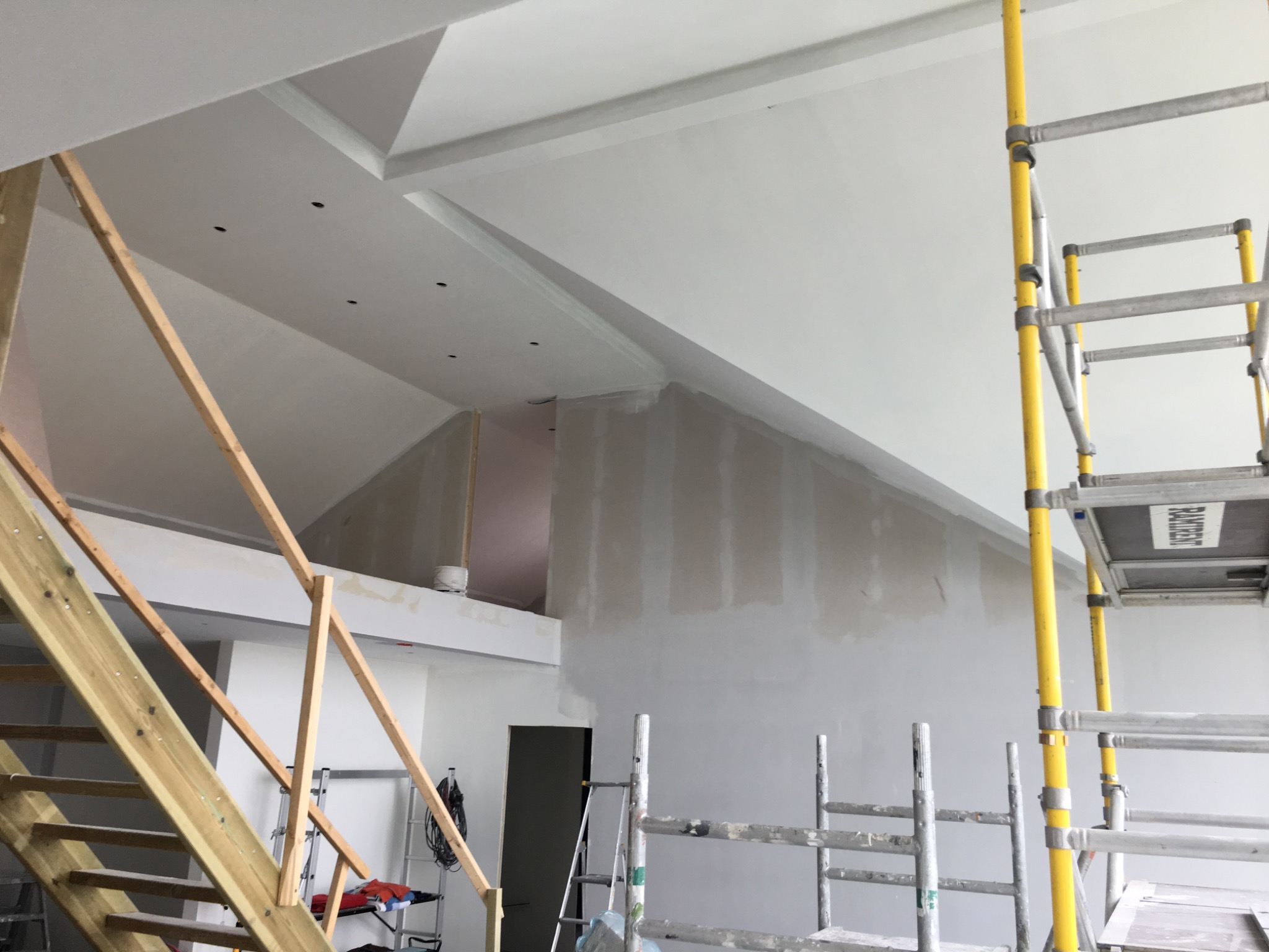 Wnętrze w trakcie prac wykończeniowych: ściany przygotowane do malowania, widoczne szpachlowanie, rusztowania, drewniane schody w surowym stanie, sufit z elementami dekoracyjnymi i punktami...