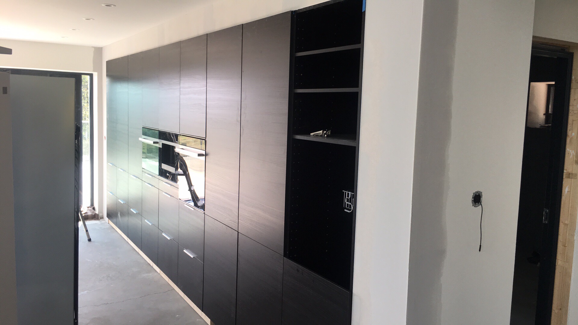 Ciemne, nowoczesne szafki kuchenne z wbudowanym piekarnikiem, otwarte półki z akcesoriami i surowa ściana z puszką elektryczną w trakcie wykańczania.