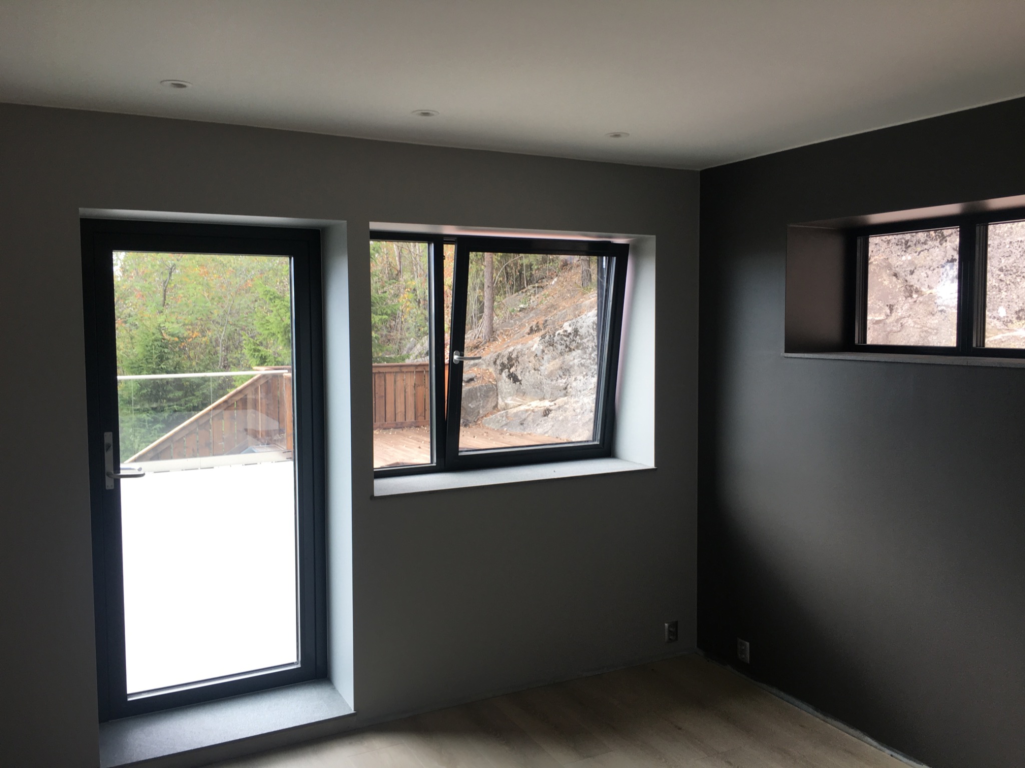 Wnętrze po remoncie: szare ściany, jasne podłogi, okno z widokiem na zewnątrz, drzwi balkonowe z białym szkłem mlecznym i małe okno w ciemnej ścianie.