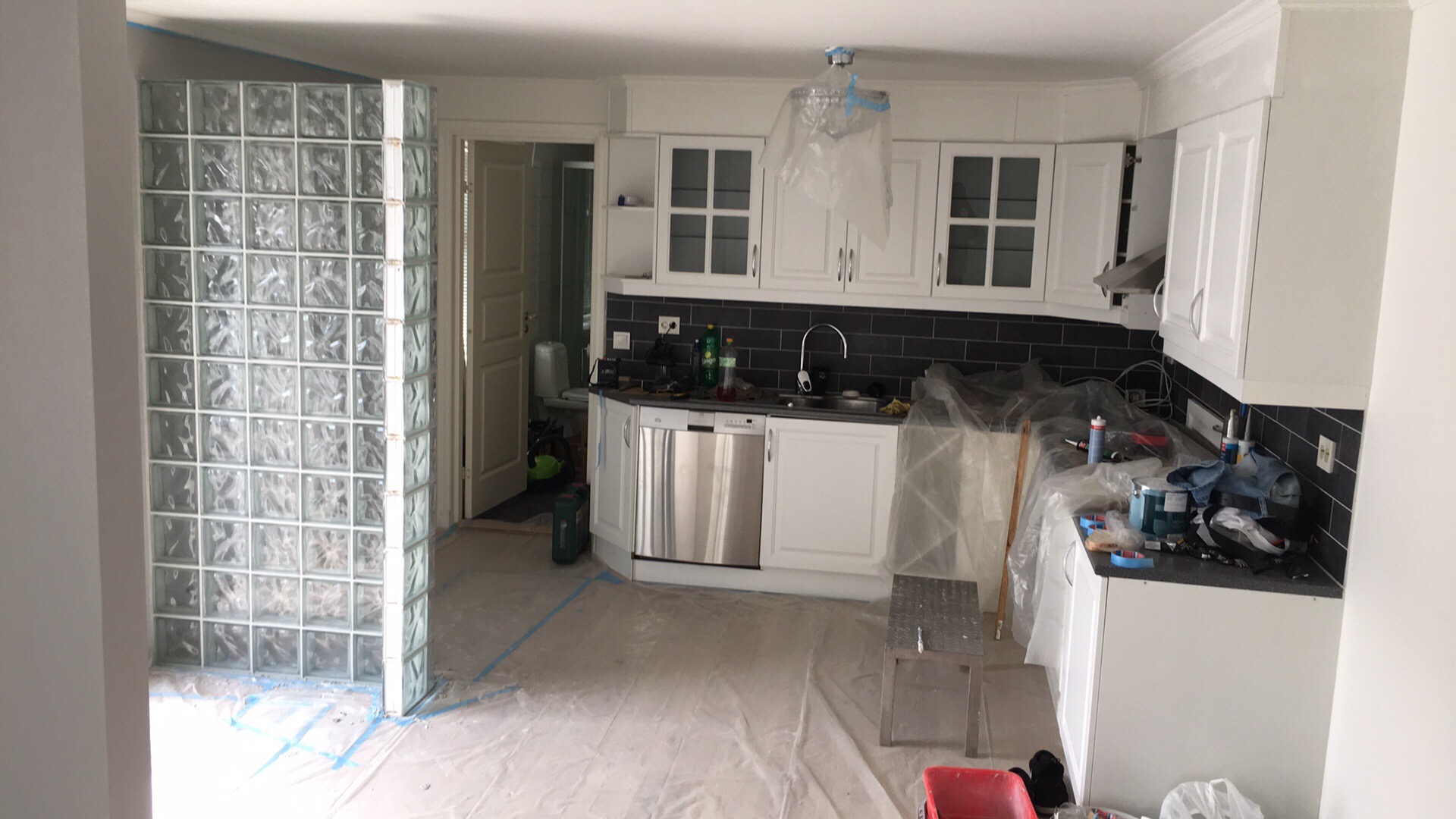 Kuchnia w trakcie remontu: ściana z luksferów, białe szafki kuchenne z czarnymi płytkami na ścianie, podłoga zabezpieczona folią malarską, widoczne narzędzia i akcesoria malarskie.