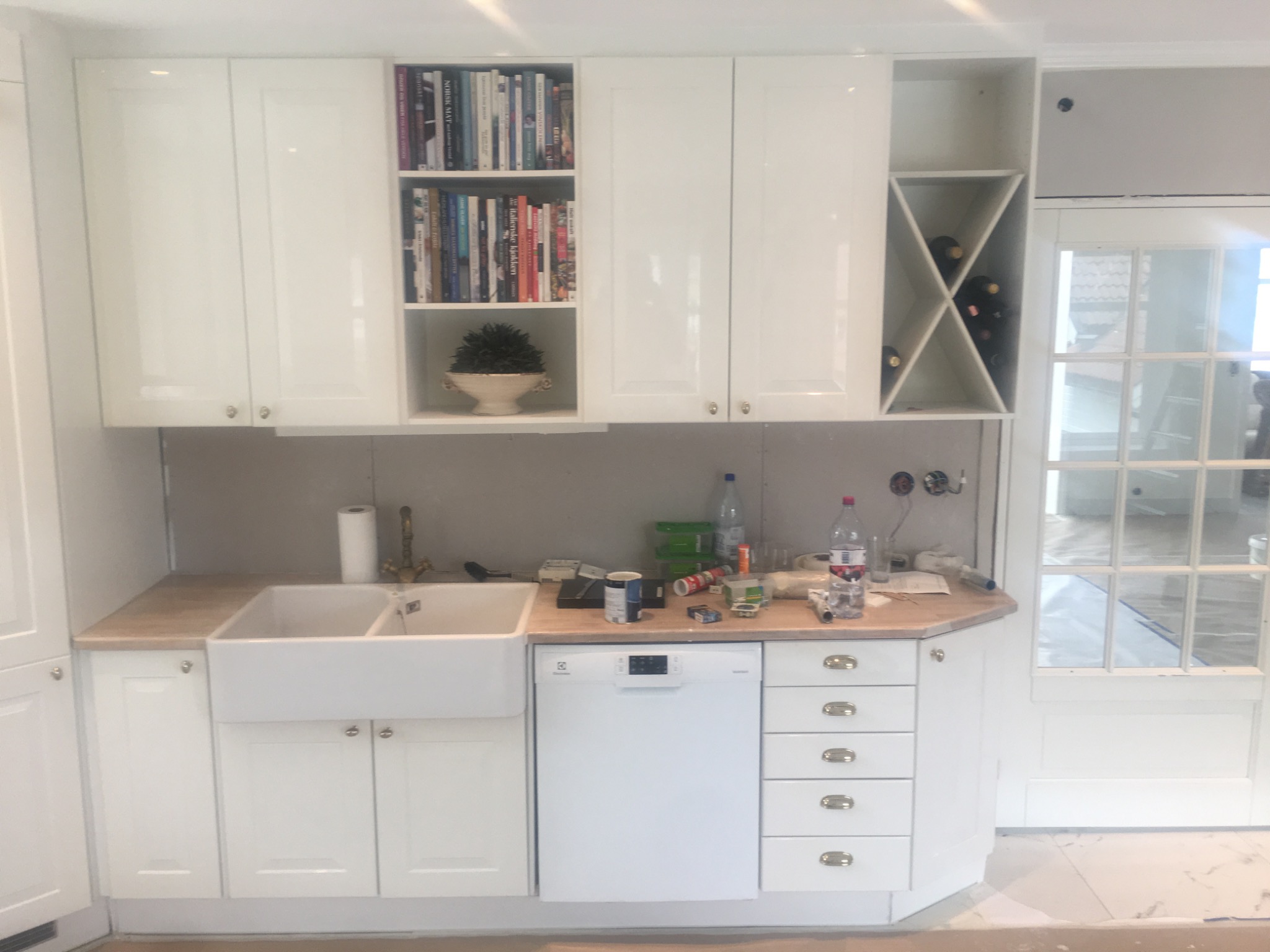 Wnętrze kuchni w trakcie prac wykończeniowych, widoczne białe szafki, zlew typu farmhouse, zmywarka, półka na książki i wino, niezabezpieczona ściana z widocznymi kablami, podłoga częściowo...