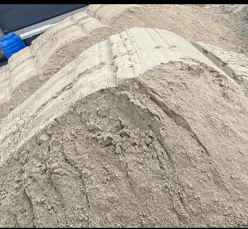 Góra jasnego piasku z widocznymi śladami opon i niebieskim pojemnikiem w tle.