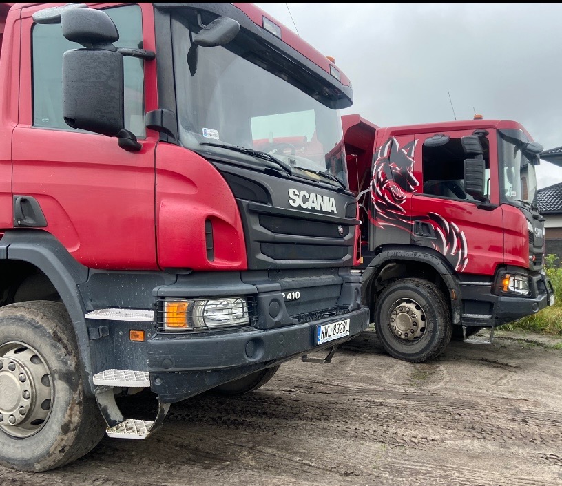 Dwa czerwone samochody ciężarowe marki Scania stoją na ziemi, jeden z grafiką wilka na kabinie, drugi z widoczną tablicą rejestracyjną.