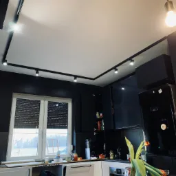 Nowoczesna kuchnia z białymi szafkami dolnymi, czarnymi szafkami górnymi i oświetleniem szynowym na suficie, widok z dołu.