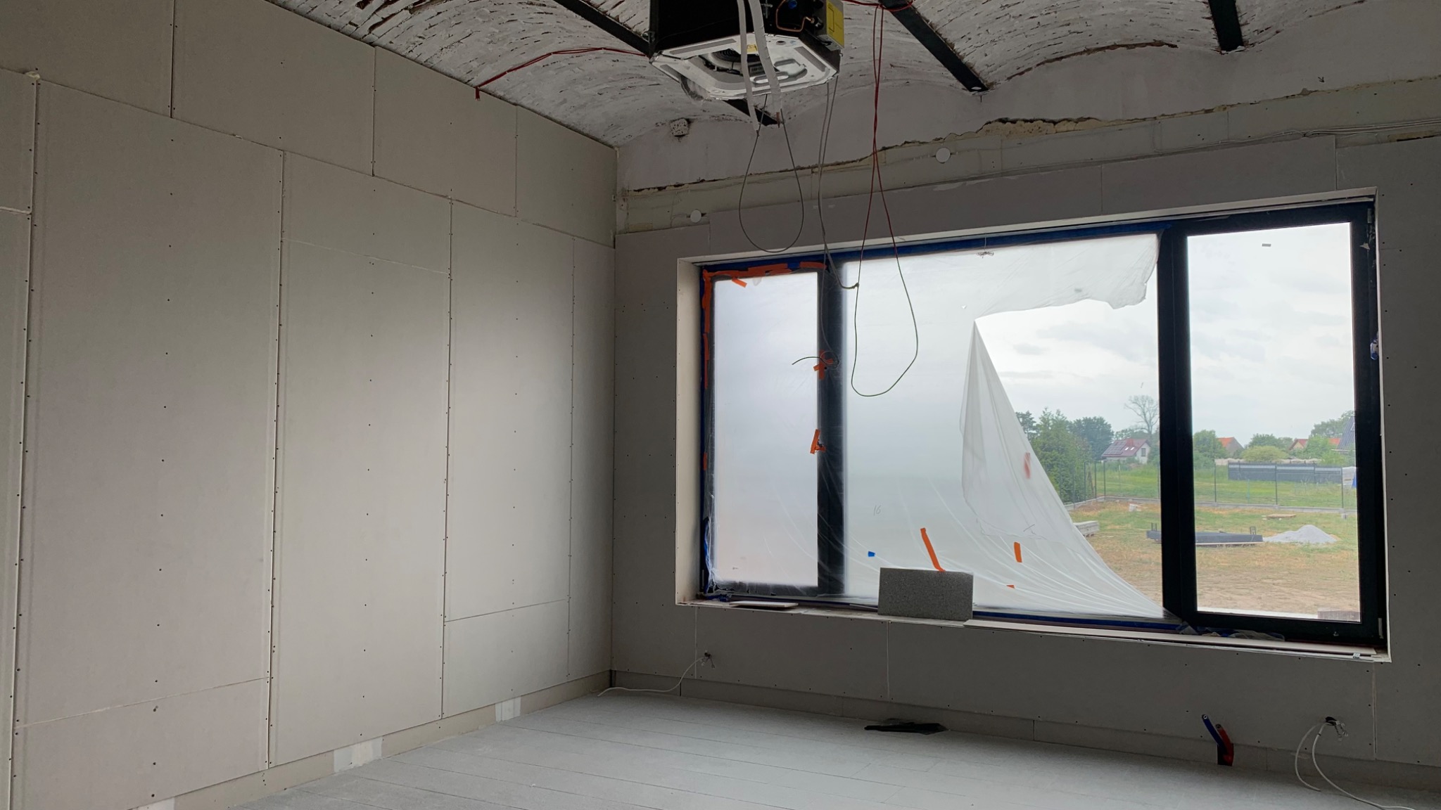 Wnętrze w trakcie prac wykończeniowych, ściany wyłożone płytami gipsowo-kartonowymi, okno zabezpieczone folią malarską, widoczna instalacja klimatyzacji pod sufitem z odsłoniętą konstrukcją.