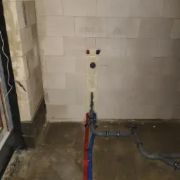 Surowy stan instalacji wodnej i kanalizacyjnej w piwnicy: czerwona i niebieska rura pionowa, szare rury kanalizacyjne z obejmami, ślady po wierceniu w ścianie z betonu komórkowego.