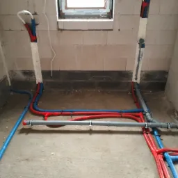 Instalacja hydrauliczna i grzewcza w surowym stanie, widoczne rury PEX w kolorach niebieskim i czerwonym, mocowane do ścian z pustaków, częściowo zabezpieczone pianką montażową.