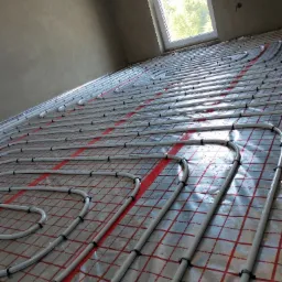 Białe rury ogrzewania podłogowego zamocowane do folii z czerwoną siatką, widok z góry w surowym wnętrzu z oknem.