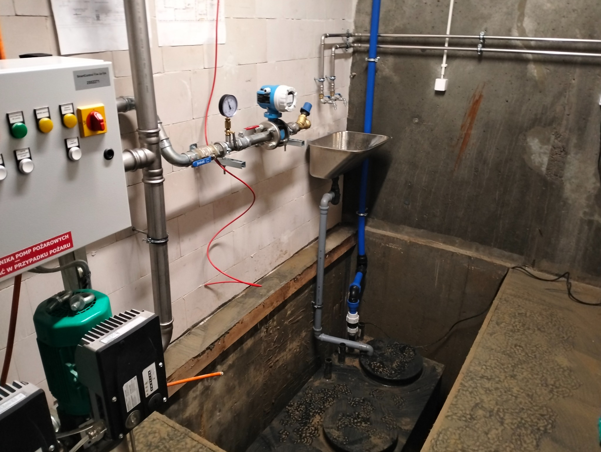 Kompleksowa instalacja pompy pożarowej w piwnicy, widoczne rury, zawory, manometr, skrzynka sterownicza z przyciskami, silnik pompy w kolorze zielonym oraz zbiornik w wykopie.