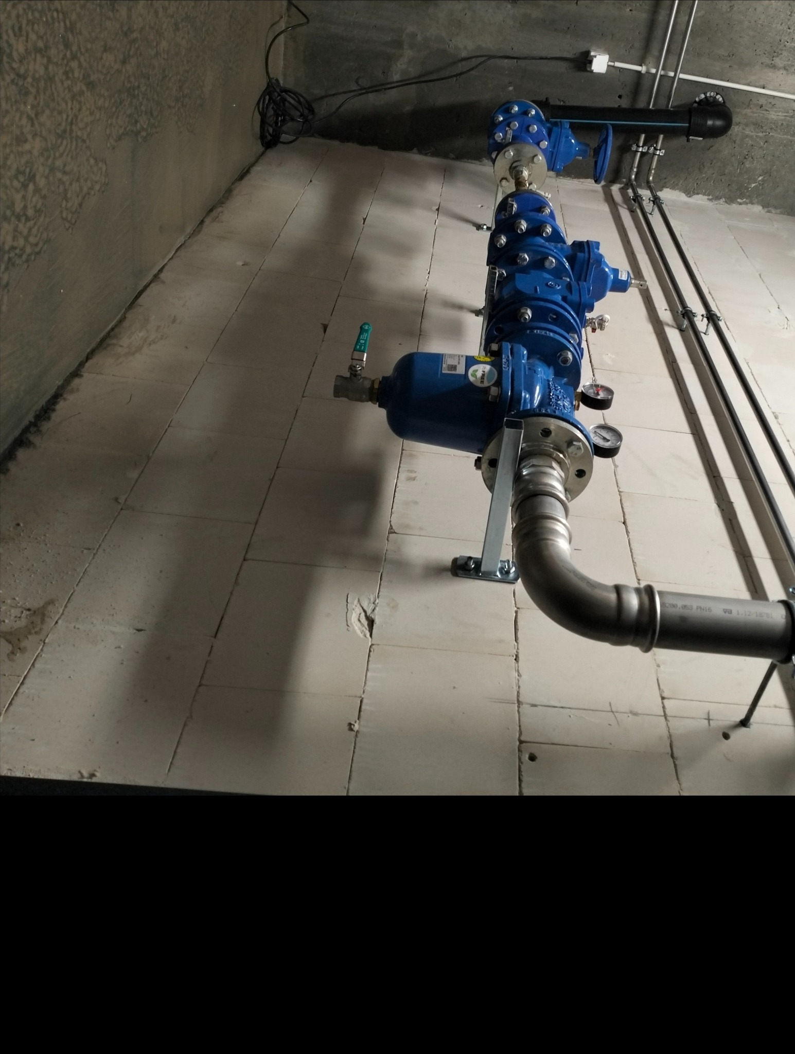 Szereg niebieskich zaworów i rur wodociągowych zamontowanych przy ścianie w piwnicy, widoczne manometry i mocowania do ściany.