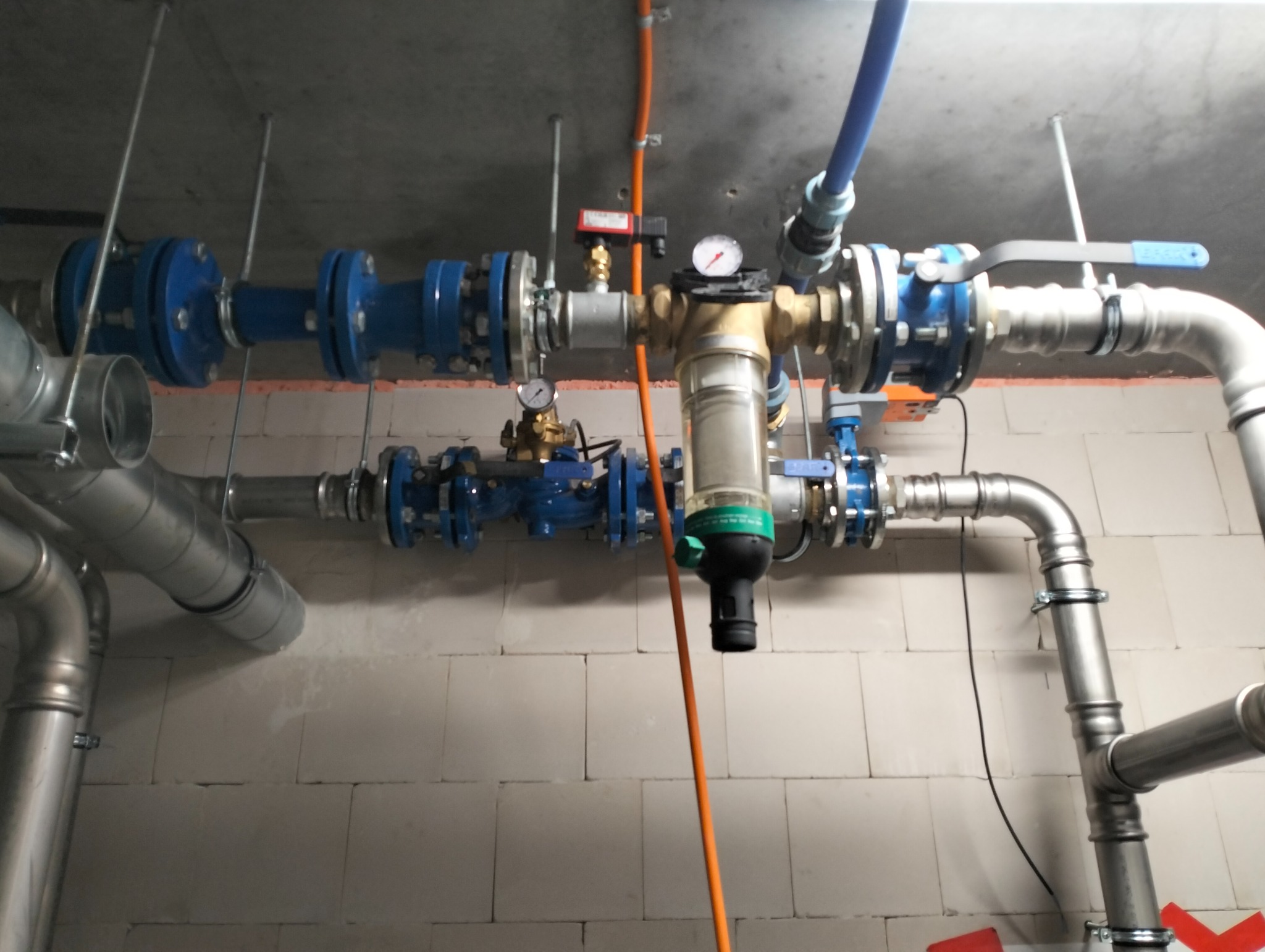 Złożony system rur hydraulicznych z zaworami, manometrami i filtrem, zamontowany pod sufitem w pomieszczeniu z surowymi ścianami z bloczków.