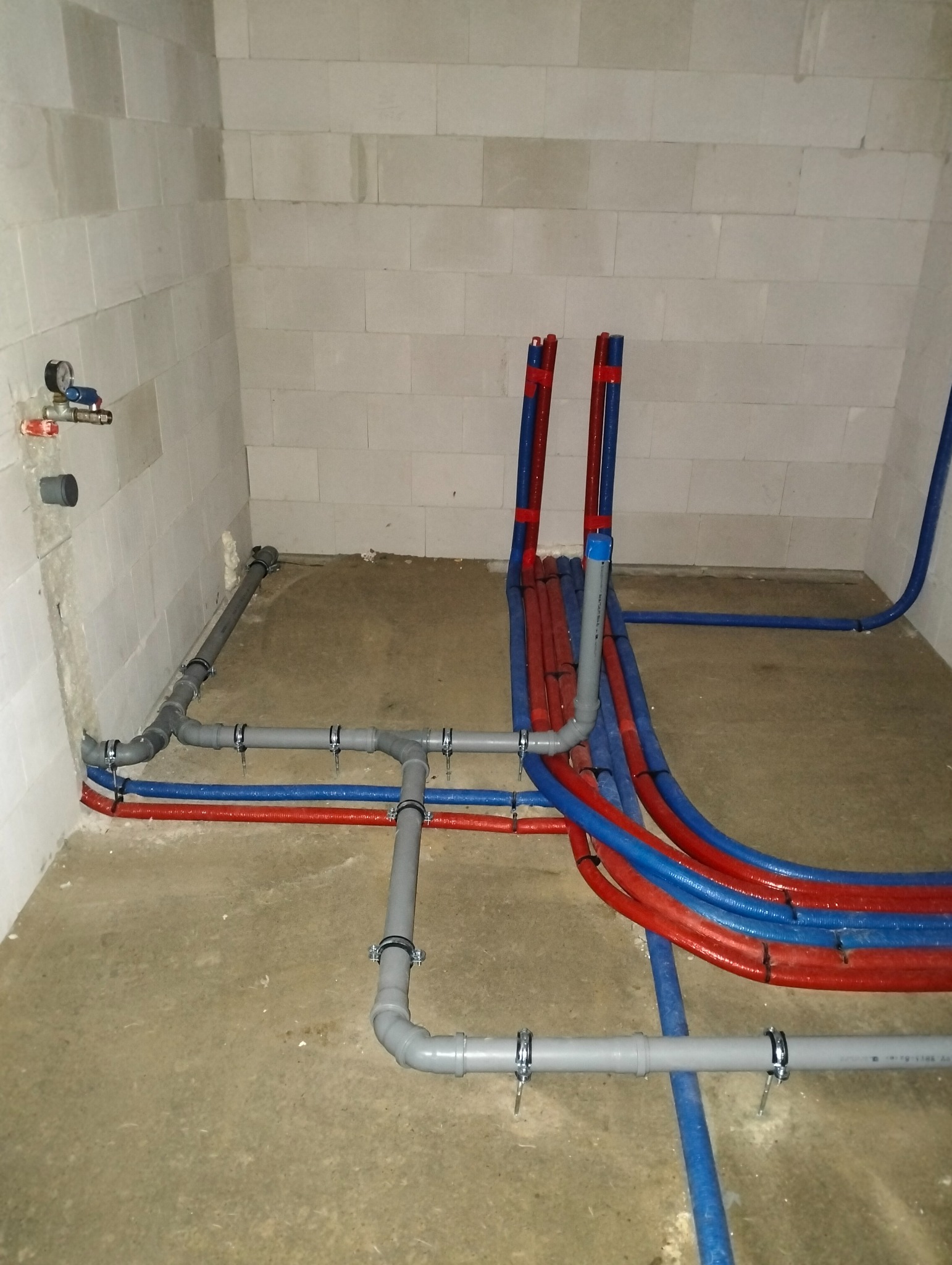 Surowe wnętrze z widocznymi rzędami czerwonych i niebieskich rur grzewczych oraz szarymi rurami kanalizacyjnymi mocowanymi metalowymi obejmami, instalacja w trakcie wykonywania.