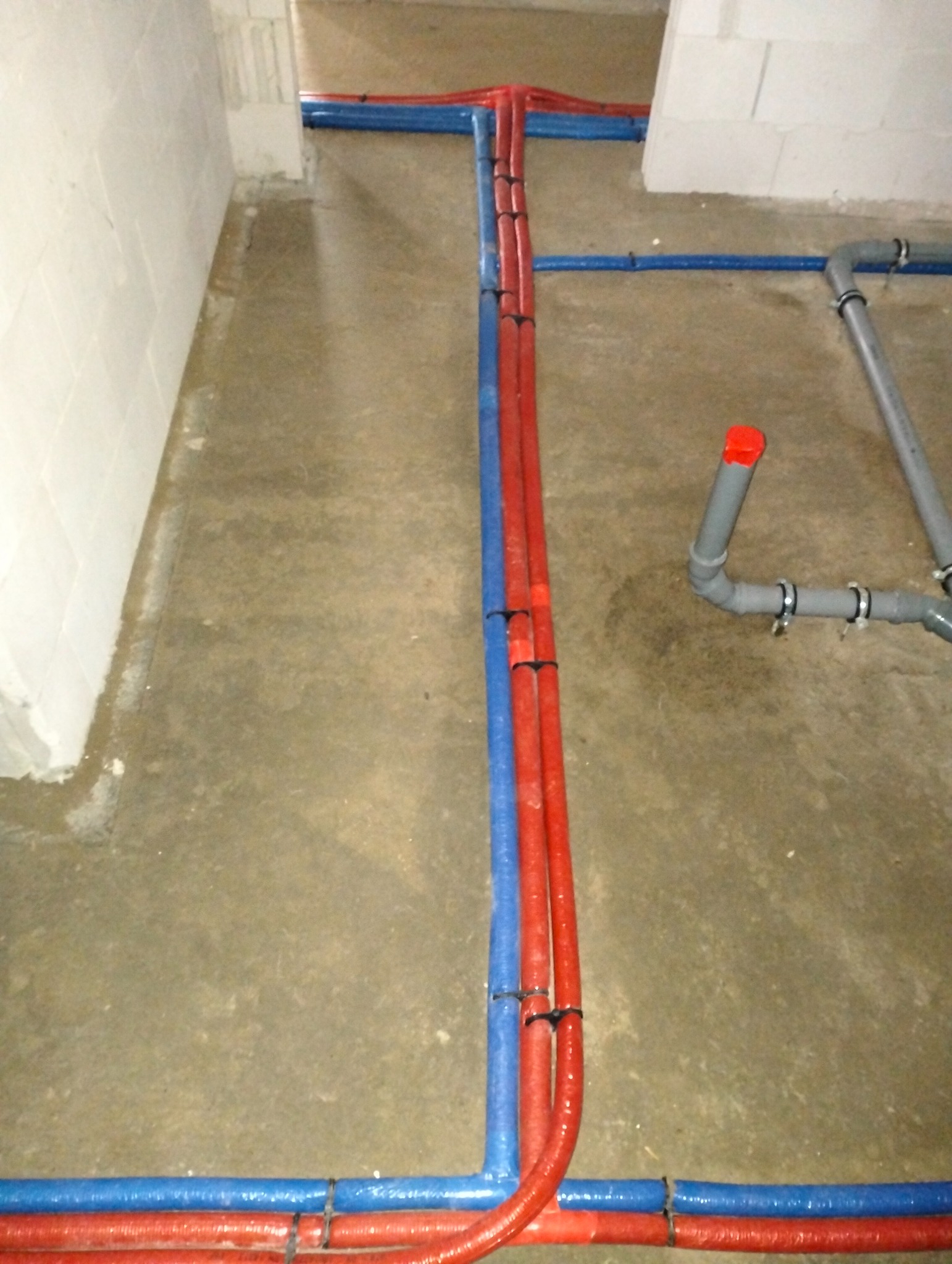 Rozprowadzenie rur instalacji grzewczej i wodnej w surowym stanie, rury w izolacji koloru czerwonego i niebieskiego, połączone czarnymi opaskami zaciskowymi, widoczne szare rury kanalizacyjne...