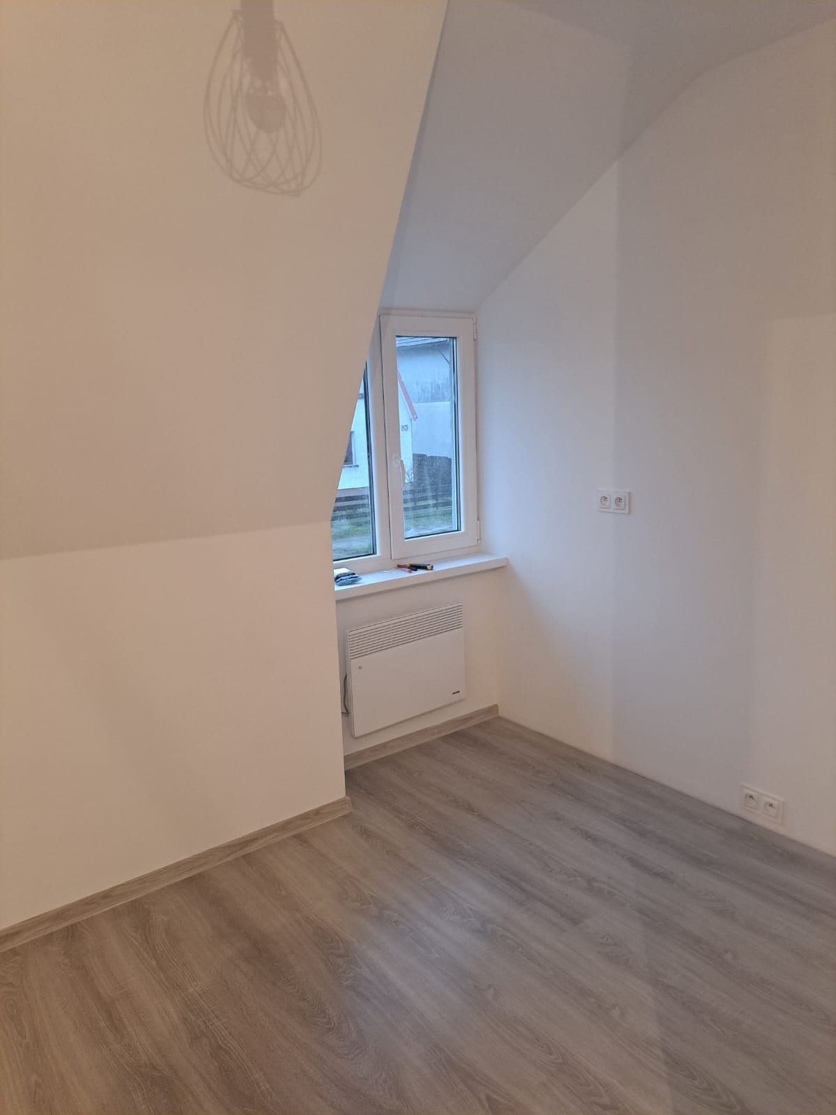 Poddasze po remoncie: świeżo pomalowane białe ściany, panelowa podłoga imitująca drewno, białe okno z widokiem na zewnątrz, grzejnik elektryczny pod oknem, cień lampy na suficie.