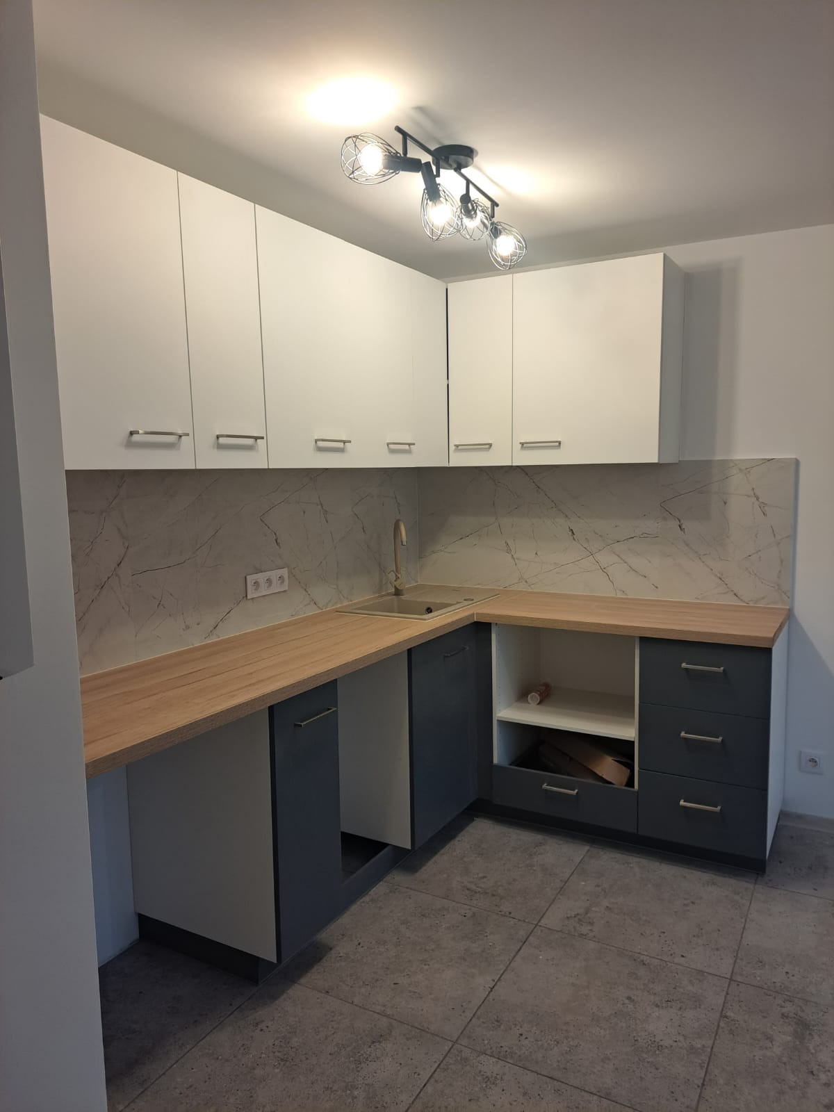 Wykończona kuchnia z białymi szafkami górnymi, szarymi dolnymi, drewnianym blatem i marmurowym panelem nad blatem. Widoczna nowoczesna lampa sufitowa i szara podłoga.