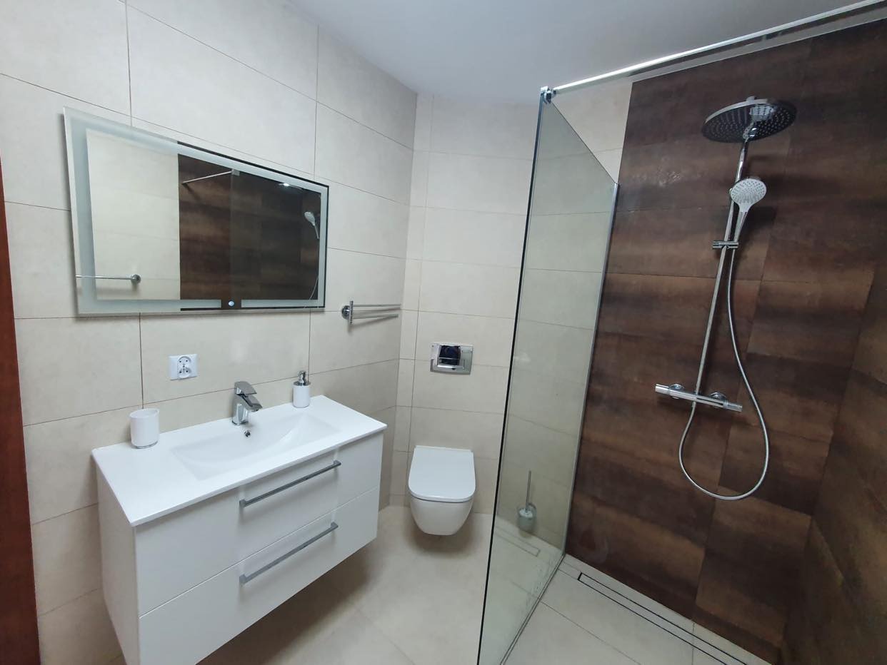 Nowoczesna łazienka z beżowymi płytkami, kabiną prysznicową typu walk-in z deszczownicą, białą umywalką z szafką i lustrem z podświetleniem LED.