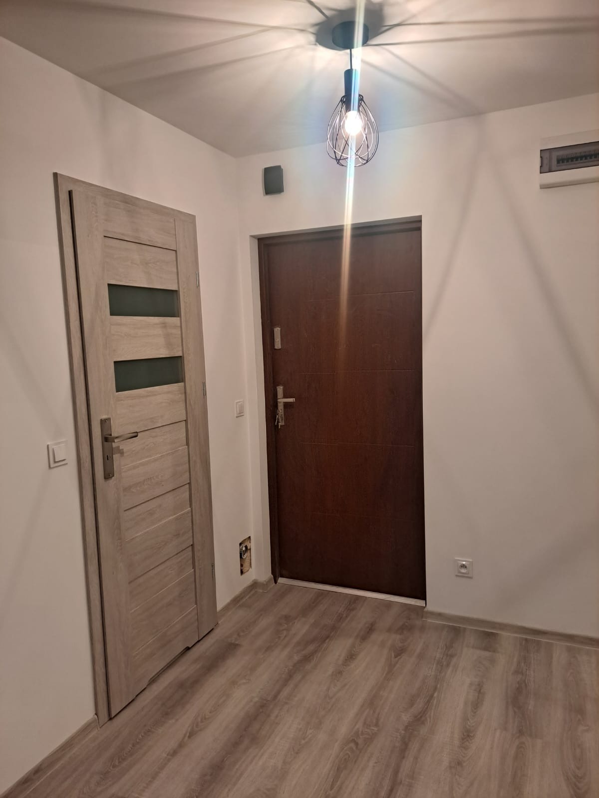 Dwie zamontowane drzwi wewnętrzne: jasne drewnopodobne z trzema przeszkleniami i ciemne, brązowe, na tle białych ścian i podłogi z paneli laminowanych.