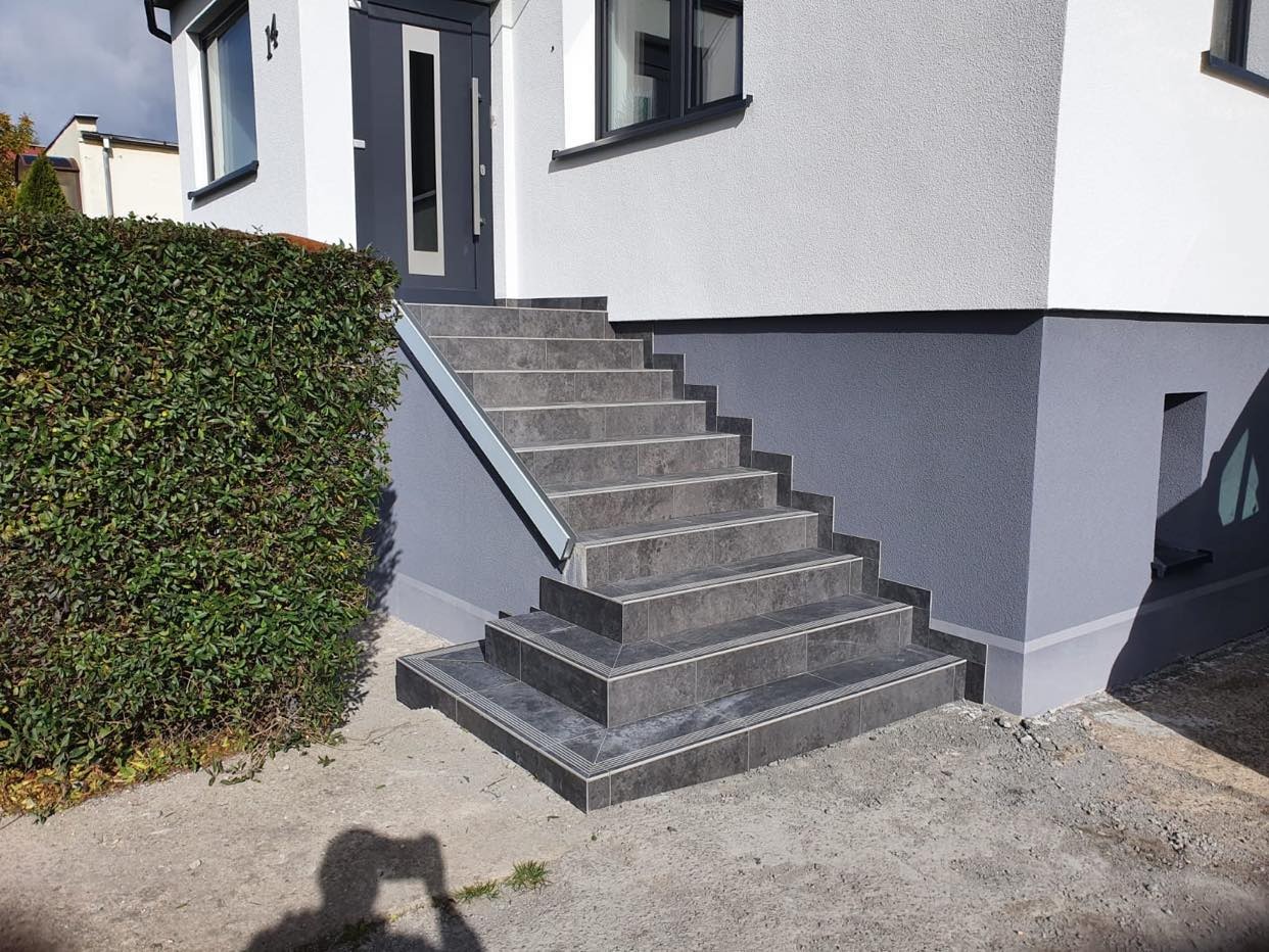 Zewnętrzne schody wejściowe do domu, wyłożone szarymi płytkami z delikatnym wzorem, z metalową listwą na krawędzi schodów, obok zadbany żywopłot