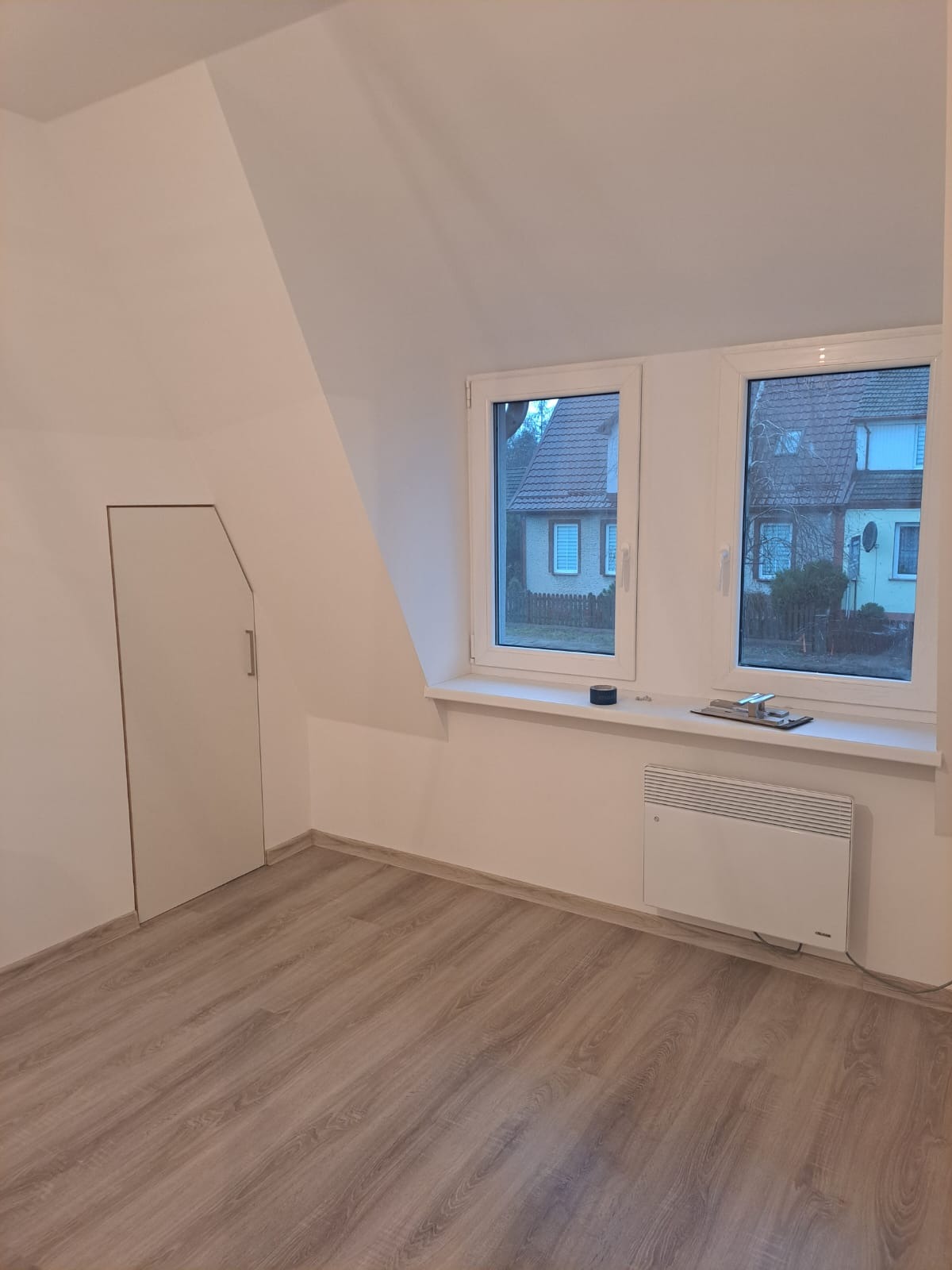 Poddasze po remoncie z drewnopodobną podłogą, białymi ścianami, grzejnikiem elektrycznym pod oknami i ukrytymi drzwiami, widok na sąsiednie domy.