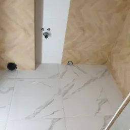 Wnętrze łazienki w trakcie prac wykończeniowych; białe płytki podłogowe z szarymi żyłami, ściany częściowo wyłożone beżowymi płytkami ułożonymi w jodełkę, widoczne instalacje hydrauliczne...