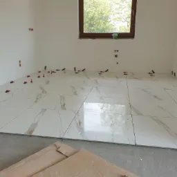 Układanie kremowych płytek podłogowych z marmurkowym wzorem w jasnym pomieszczeniu, widoczne plastikowe krzyżyki dystansowe, okno w drewnianej ramie i niezakończone prace wykończeniowe.