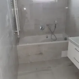 Nowoczesna łazienka z wanną obłożoną płytkami imitującymi marmur, grzejnikiem drabinkowym i białymi szafkami pod umywalką, oświetlona naturalnym światłem z okna.