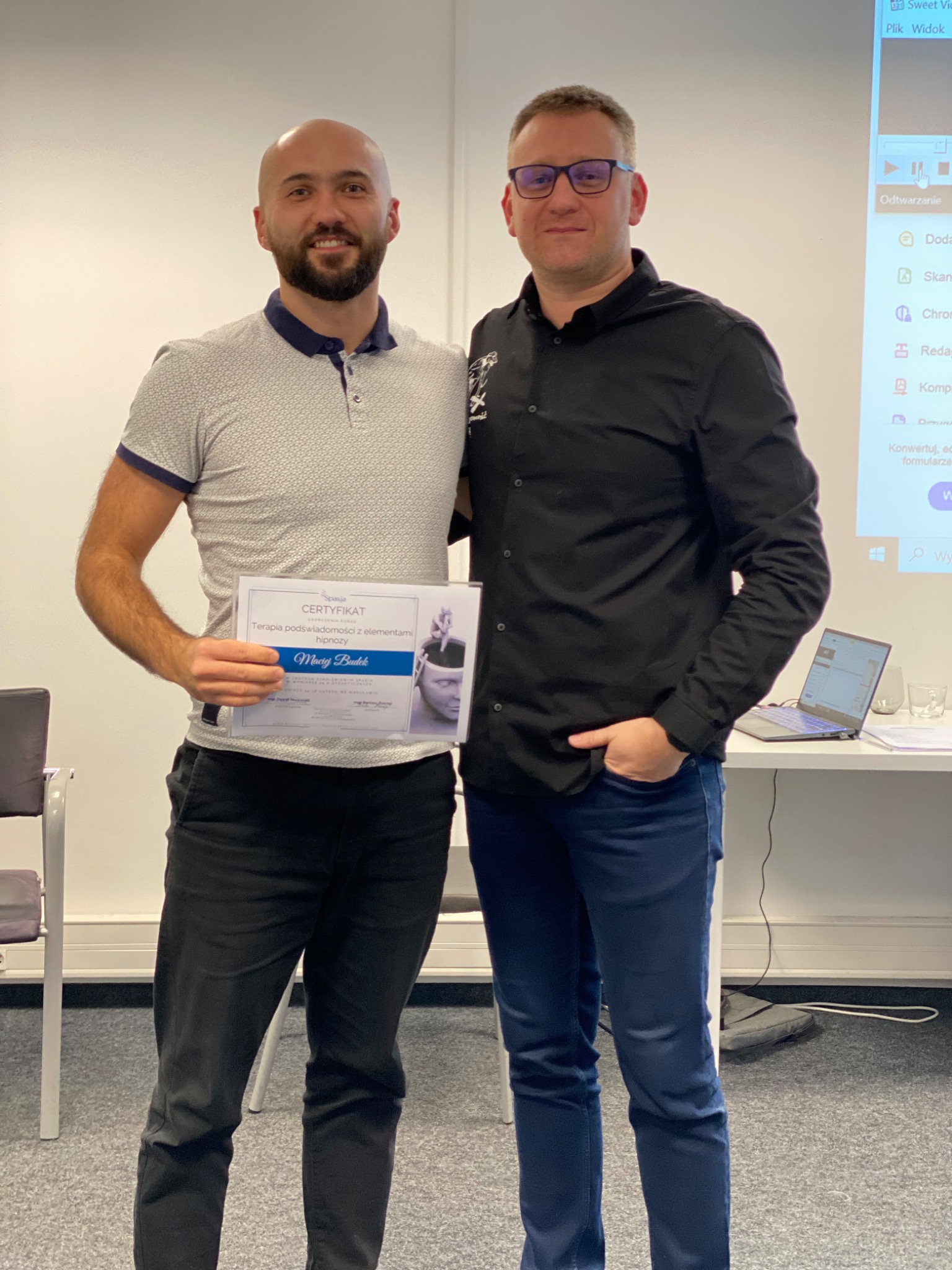 Dwóch mężczyzn pozuje z certyfikatem ukończenia kursu terapii podświadomości z elementami hipnozy, trzymanym przez jednego z nich. W tle laptop i projektor.