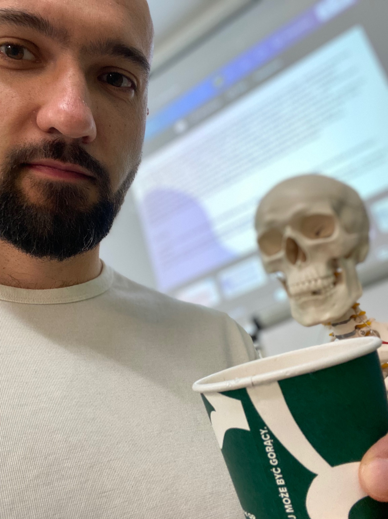 Mężczyzna z brodą i kubkiem, obok szkielet. W tle ekran z prezentacją. Nietypowe ujęcie w gabinecie fizjoterapii lub podczas zajęć z anatomii.