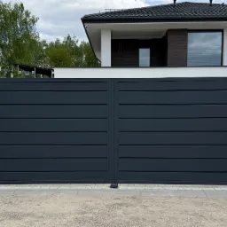 Nowoczesna, przesuwna brama wjazdowa w kolorze grafitowym, z poziomymi panelami, osadzona na murowanych słupkach z jasnego kamienia. W tle dom z ciemnym dachem.