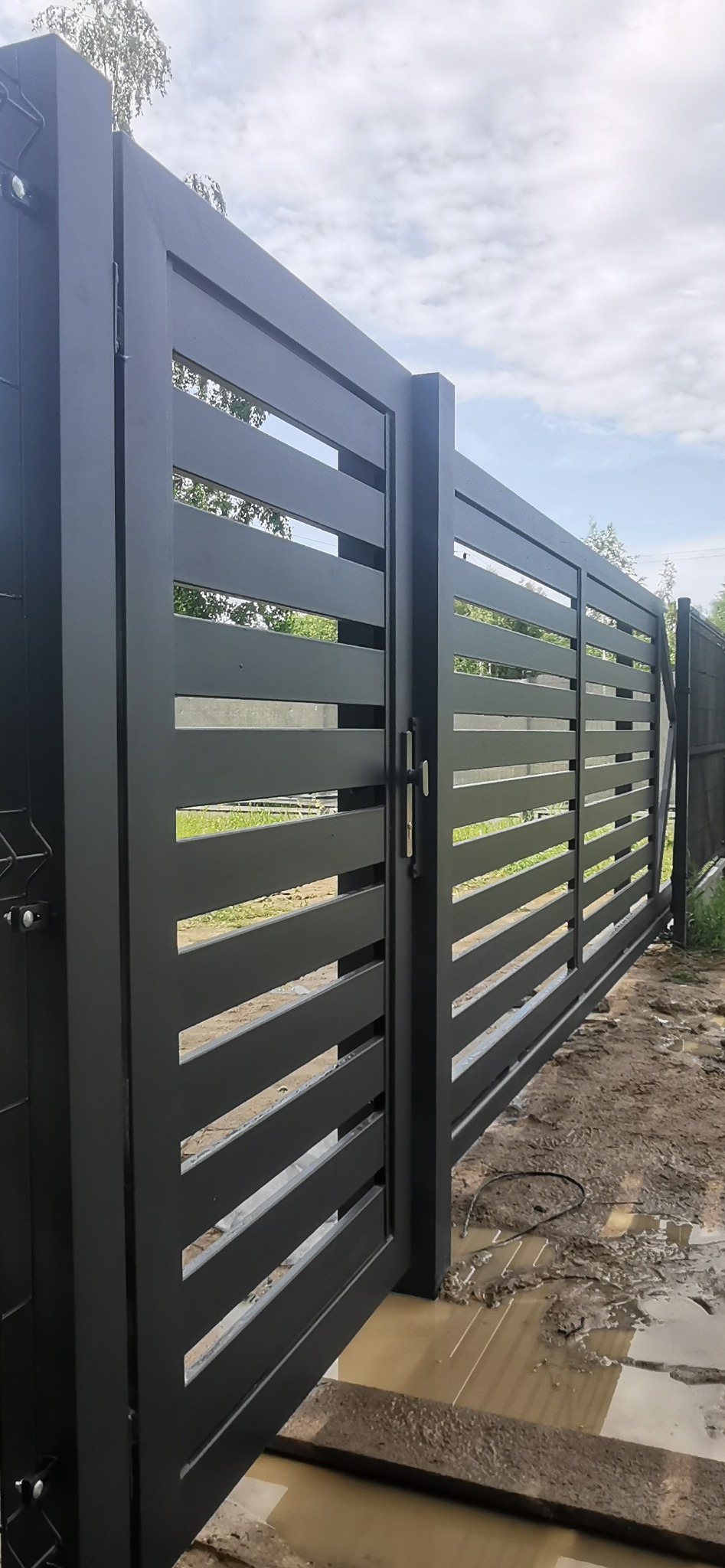 Nowoczesna, otwarta brama wjazdowa z poziomymi panelami, pomalowana na czarno, z widocznym fragmentem ogrodzenia panelowego w tle, na posesji w trakcie prac ziemnych.