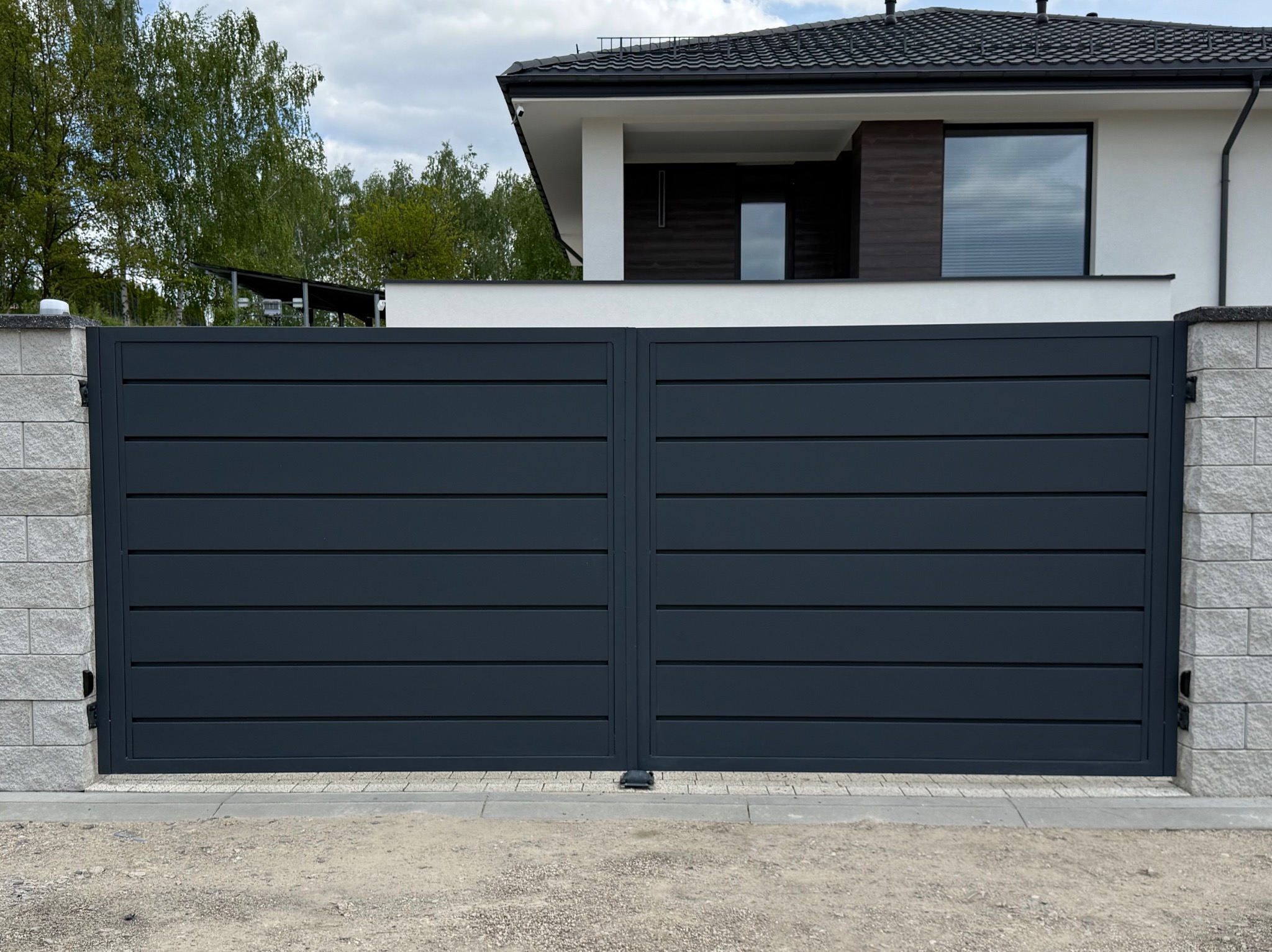 Nowoczesna, przesuwna brama wjazdowa w kolorze grafitowym, z poziomymi panelami, osadzona na murowanych słupkach z jasnego kamienia. W tle dom z ciemnym dachem.