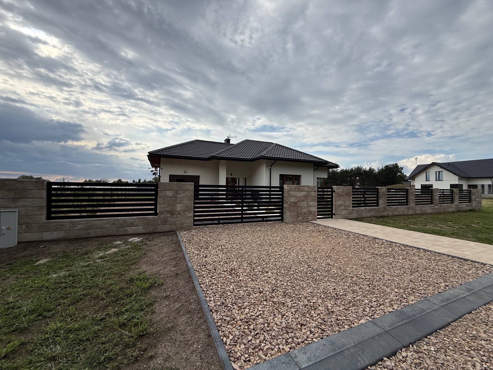 Nowoczesne ogrodzenie z poziomych, czarnych paneli i betonowych słupków, otaczające posesję z domem jednorodzinnym. Podjazd wysypany drobnym kamieniem.
