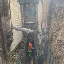 PM SEWERS - Głęboki wykop zabezpieczony stalowymi szalunkami, wewnątrz pracownik w odblaskowej odzieży łączy pomarańczową rurę; na górze widoczny fragment koparki.