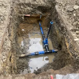 Wykop z ułożonymi rurami wodociągowymi i kanalizacyjnymi wypełniony wodą, widoczne narzędzia pracy na brzegu wykopu.