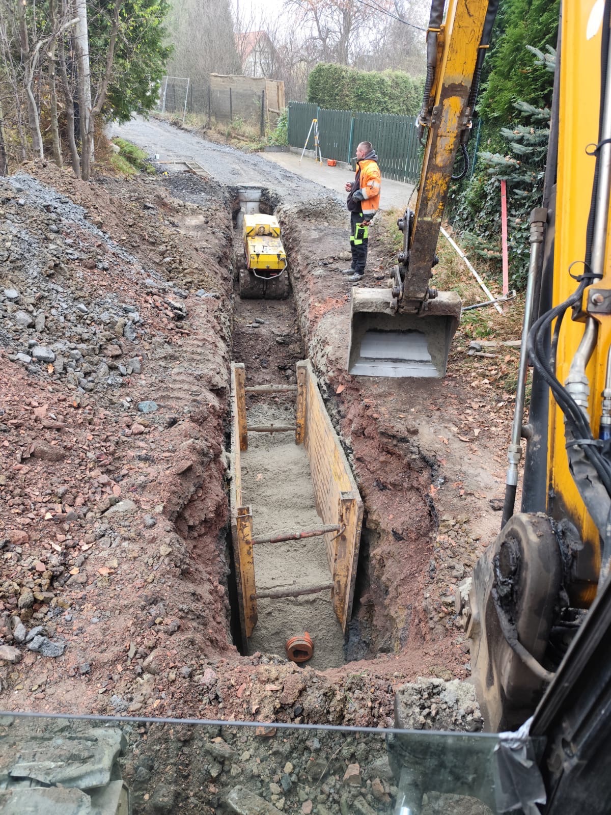 Głęboki wykop zabezpieczony szalunkiem stalowym, wewnątrz rura kanalizacyjna, na górze ubijarka i operator w odblaskowej kamizelce, na pierwszym planie ramię koparki, teren budowy.