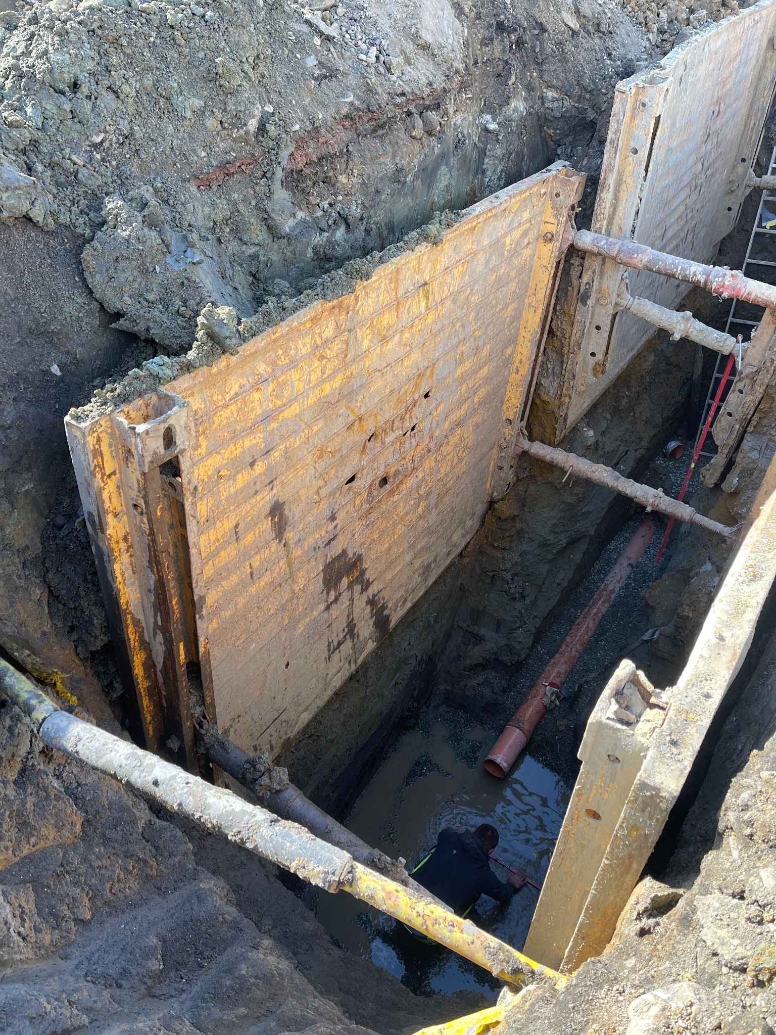 Głęboki wykop zabezpieczony metalowymi szalunkami, w dole pracownik w wodzie, widoczne rury instalacyjne i drabina, ziemia wokół wykopu.